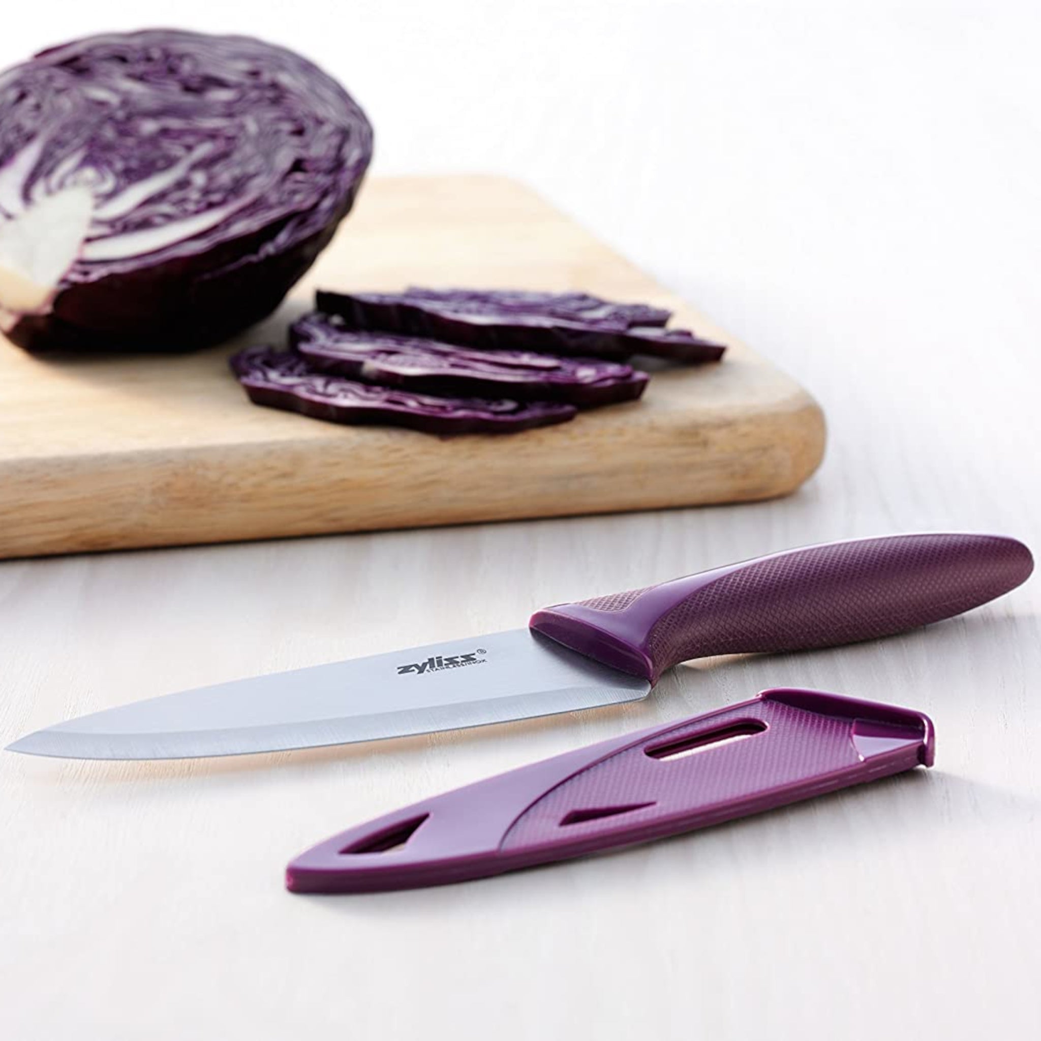 A Zyliss 3 Piece Knife Set chef’s knife with a purple handle rests on a light wooden surface beside slices of red cabbage, highlighting one of the most versatile and essential kitchen tools.