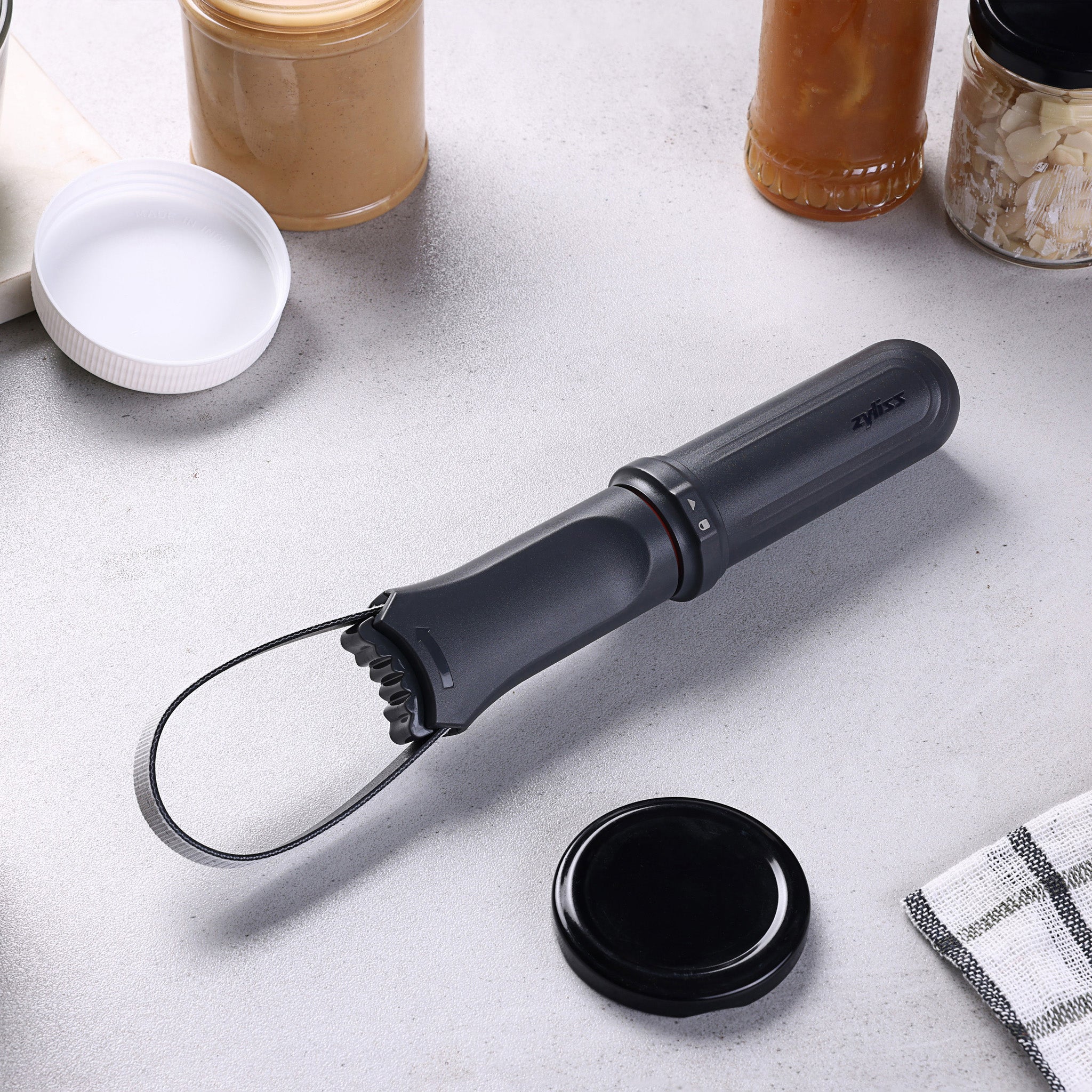 A black electric jar opener with a rubber strap lies on a kitchen counter surrounded by jars of peanut butter, honey, and garlic, along with a dish towel and jar lids.
