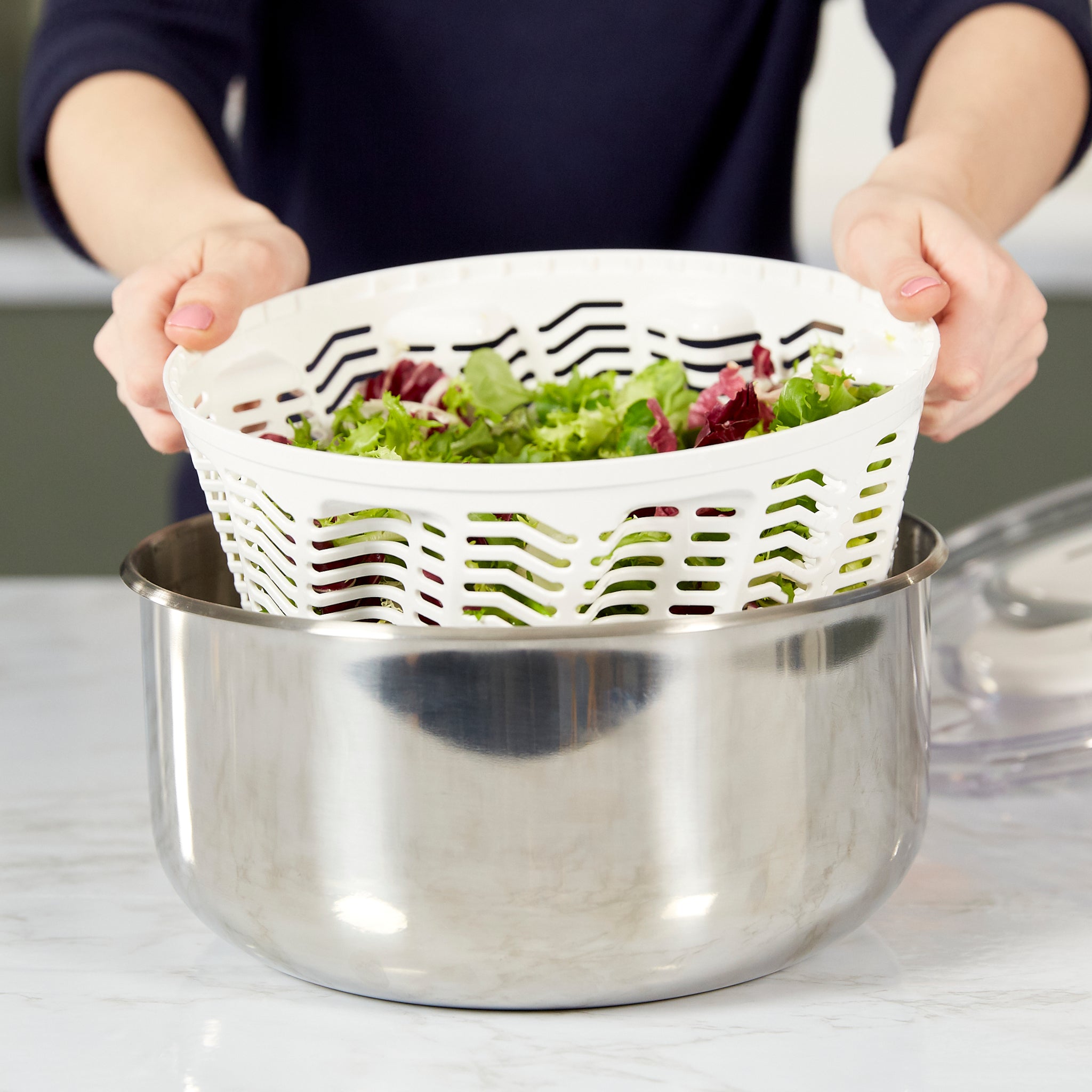 A person using a salad spinner, lifting the perforated basket filled with leafy greens over a stainless steel bowl on a kitchen counter.