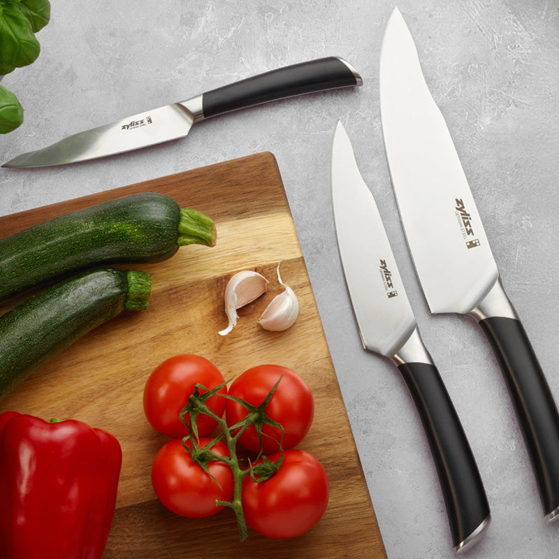 Three kitchen knives are placed on a gray surface next to a wooden cutting board holding two zucchinis, a red bell pepper, garlic cloves, and vine tomatoes.