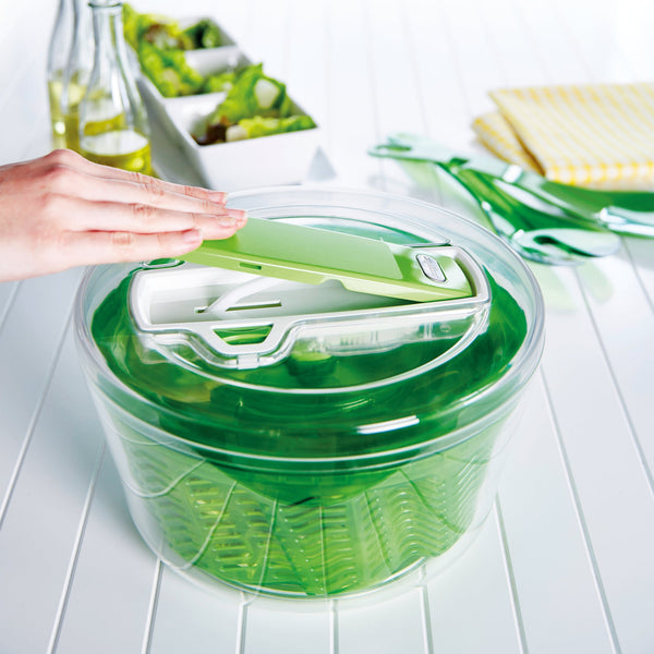 A person uses the Zyliss Swift Dry Salad Spinner Large, featuring a green and white design, clear bowl, and non-slip base on a white table. Green utensils, a yellow-striped napkin, bottles, and salad dishes are visible in the background.