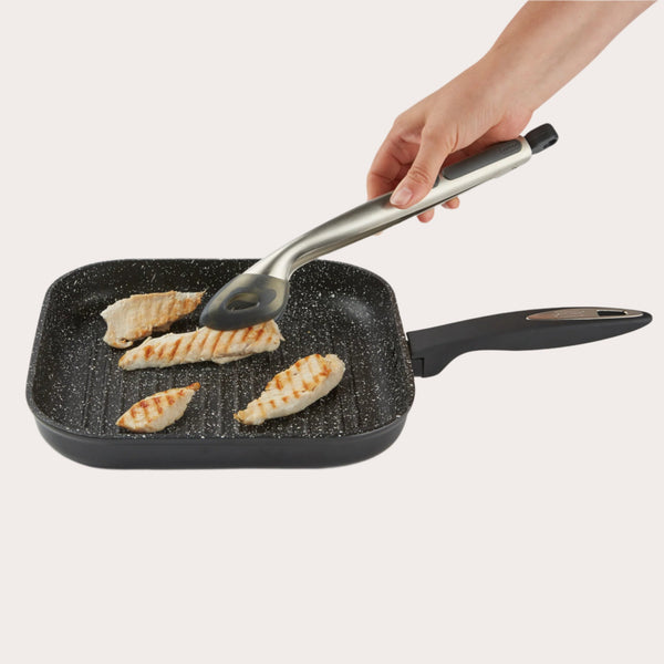 A hand uses Zyliss Silicone-Tipped Tongs to turn grilled chicken breast in a black speckled non-stick grill pan with a handle, set against a plain white background.