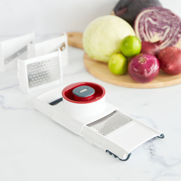 A Zyliss 4-in-1 Slicer & Grater in white and red sits on a kitchen countertop, with cabbage, limes, onions, and a beet arranged on a wooden board in the background. 