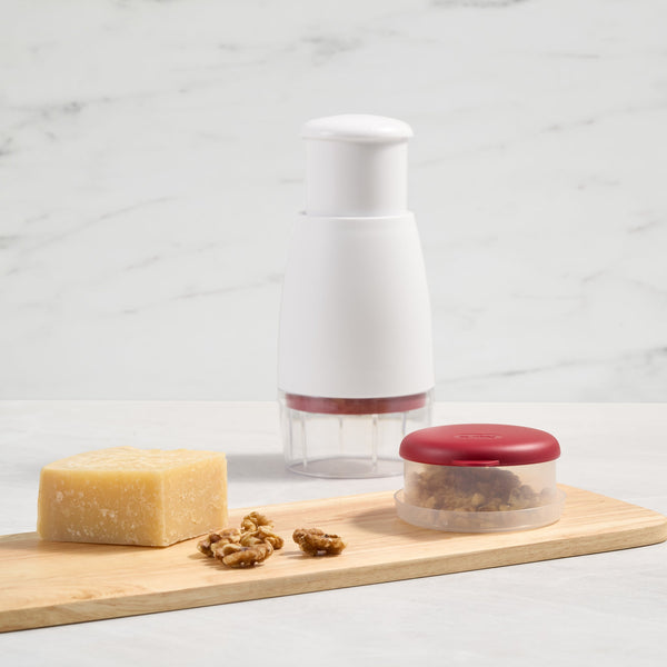 A Zyliss Zick-Zick Classic Food Chopper sits on a marble surface behind a wooden board holding cheese, walnuts, and a small red-lidded storage container filled with chopped nuts. 