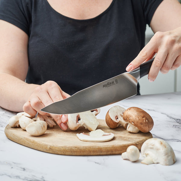 Wearing a black shirt, a person slices white and brown mushrooms on a wooden board with the Zyliss Comfort Pro Chefs Knife 20cm, featuring an ergonomic handle and precision German stainless steel blade on a marble countertop. 