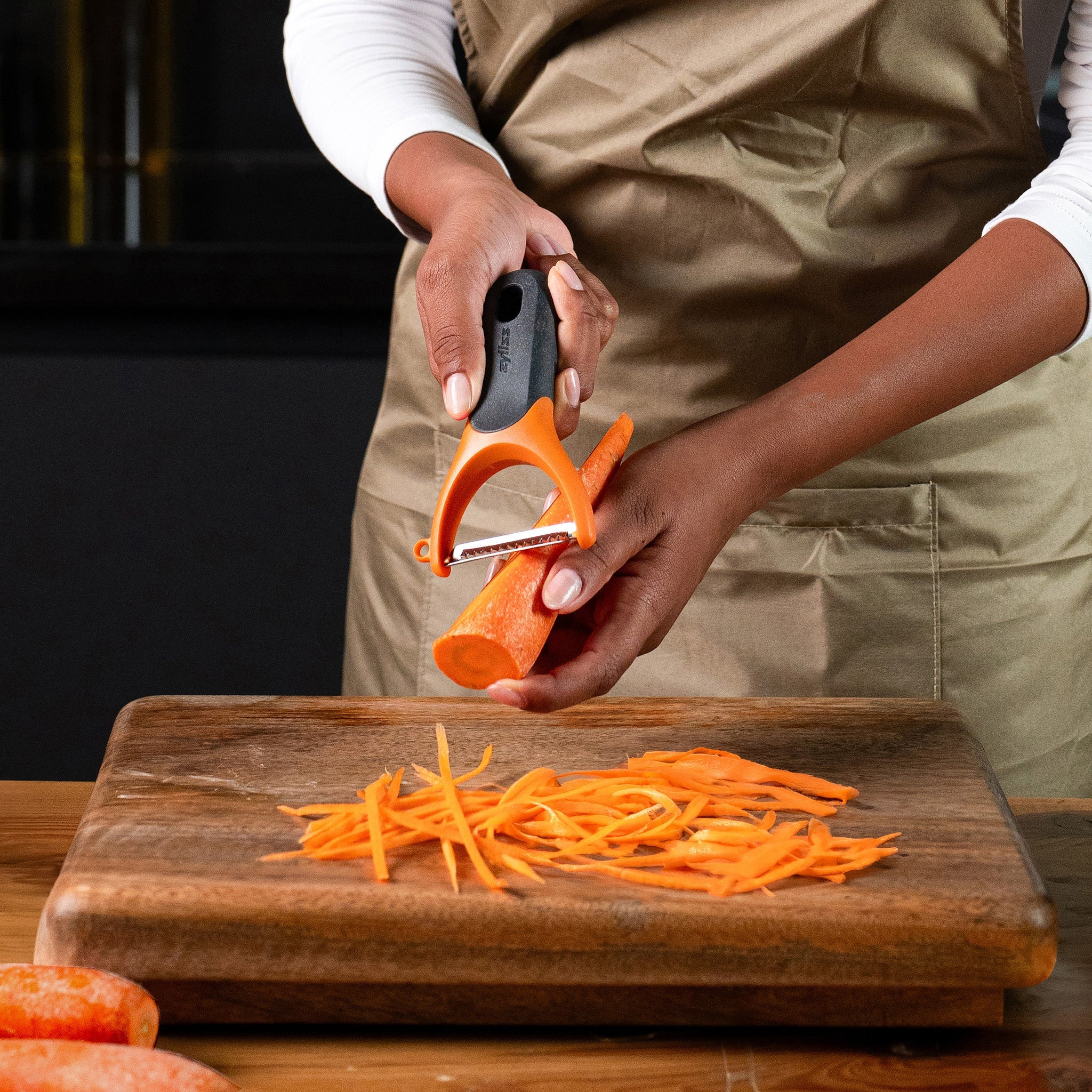 A person wearing a beige apron uses a vegetable peeler to julienne a carrot over a wooden cutting board, with thin carrot strips scattered on the board.