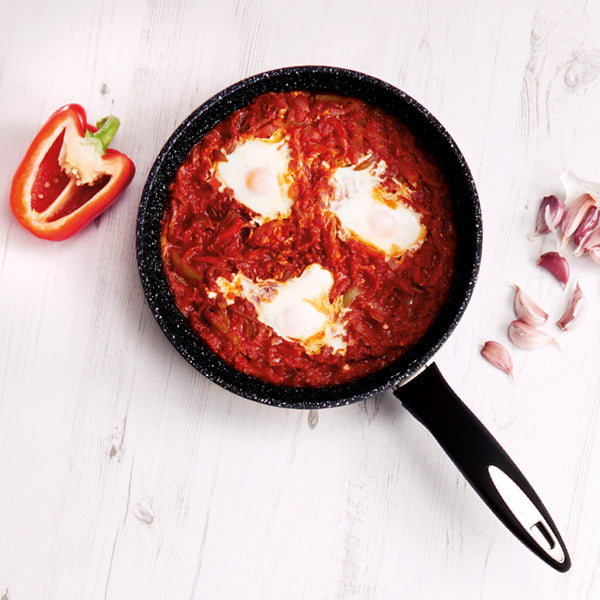 A black frying pan with shakshuka—tomato sauce and three eggs—on a white surface. Nearby are a halved red bell pepper and several garlic cloves.