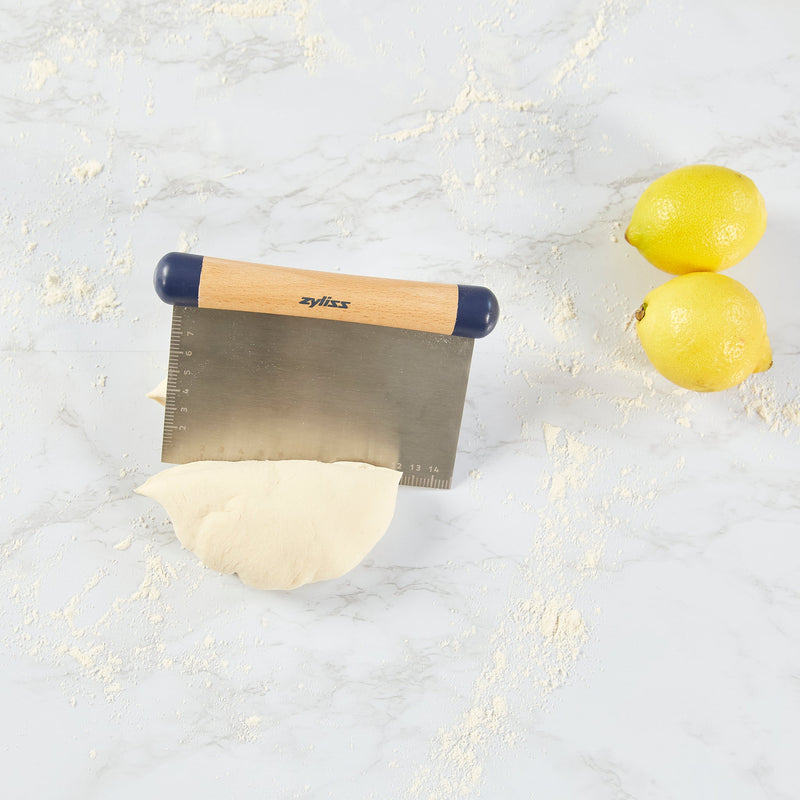 The Zyliss Beech Wood Bench Scraper, a versatile baking tool, slices through dough on a floured surface with two lemons nearby. 
