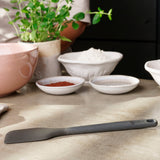 A black Zyliss Spatula with a silicone head rests on a kitchen counter near bowls of flour, cocoa, and sugar. In the background, someone stands by a pink mixing bowl and a potted herb plant. #Medium