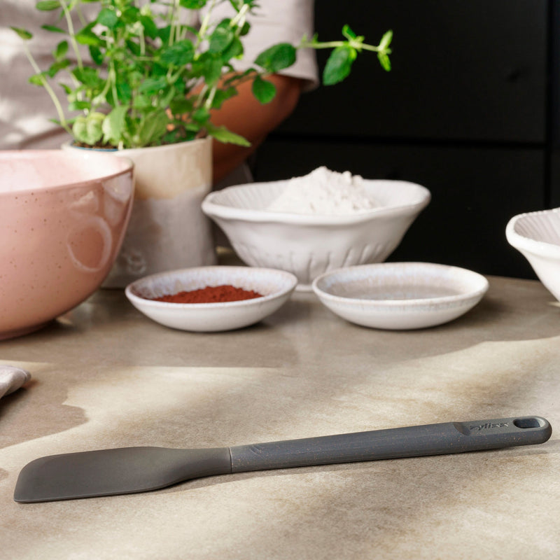 A black Zyliss Spatula with a silicone head rests on a kitchen counter near bowls of flour, cocoa, and sugar. In the background, someone stands by a pink mixing bowl and a potted herb plant. #Medium