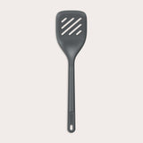 The Zyliss Slotted Turner, shown in black with three diagonal slots and an ergonomic handle, features a heat-resistant design for safe cooking or flipping food. Displayed against a plain white background. 