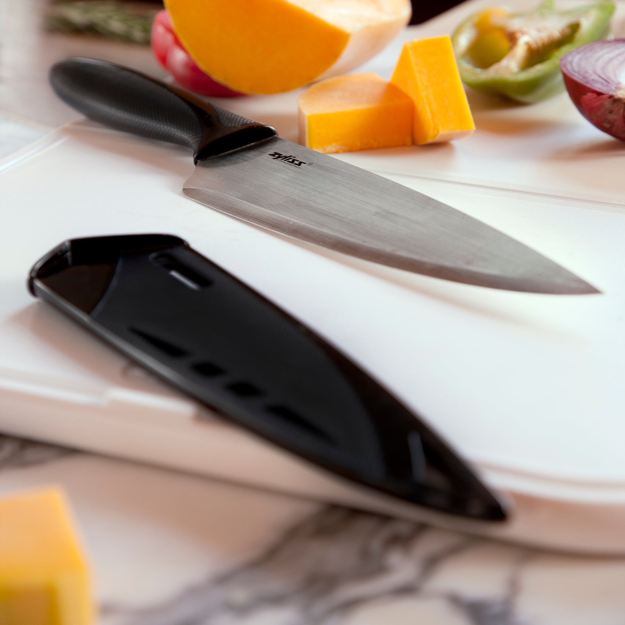 A sharp kitchen knife with a black handle rests on a white cutting board beside its black sheath. Sliced vegetables, including onion, bell pepper, and cheese, are scattered around on a marble countertop.
