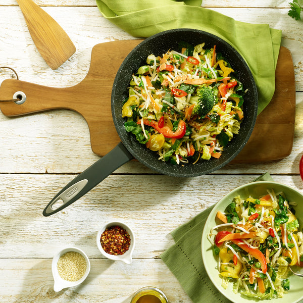 The Zyliss Ultimate Non-Stick Frying Pan, filled with vibrant stir-fried veggies, rests on a wooden board beside a matching bowl. Nearby, small bowls of sesame seeds and chili flakes sit on a rustic white table. #2Pc_Set
